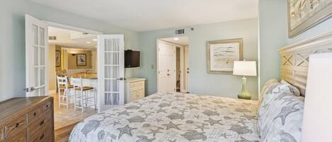 A bedroom with a bed, dresser, and a dining area visible through an open door.