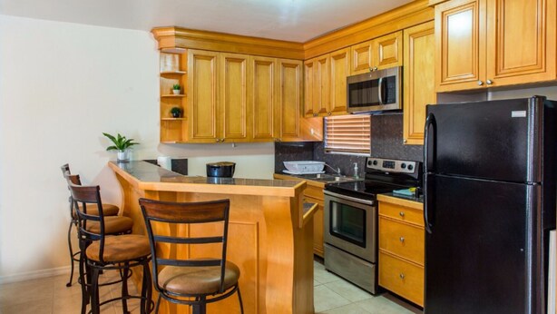 A kitchen with wooden cabinets, a black refrigerator, a microwave, an oven, and a sink. There are two bar stools and a potted plant. - Caicos Cactus Residence  Major Hill (Caicos Islands)