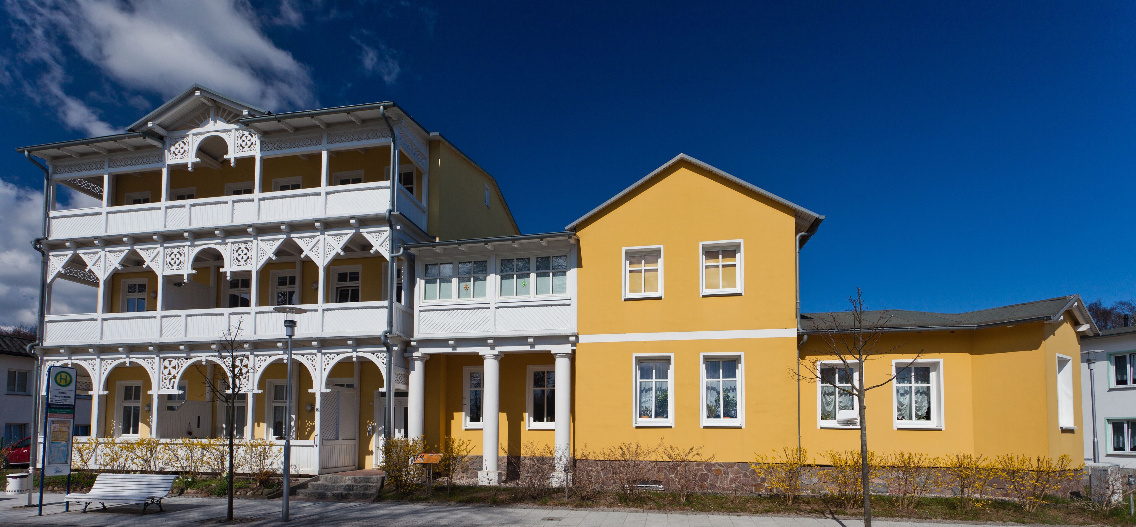 A two-story building with a yellow facade and white trim, featuring a balcony with intricate lattice work.