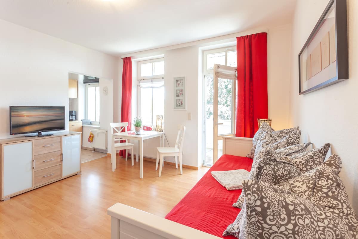 A modern living room with a sofa, a TV, a dining area with a table and chairs, and large windows with red curtains.