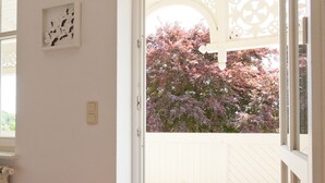 A room with a wooden floor, a red and white striped rug, a blue doormat, and a white door leading to an outdoor area with a wooden deck and flowering trees. - Haus Sonne (Sellin)