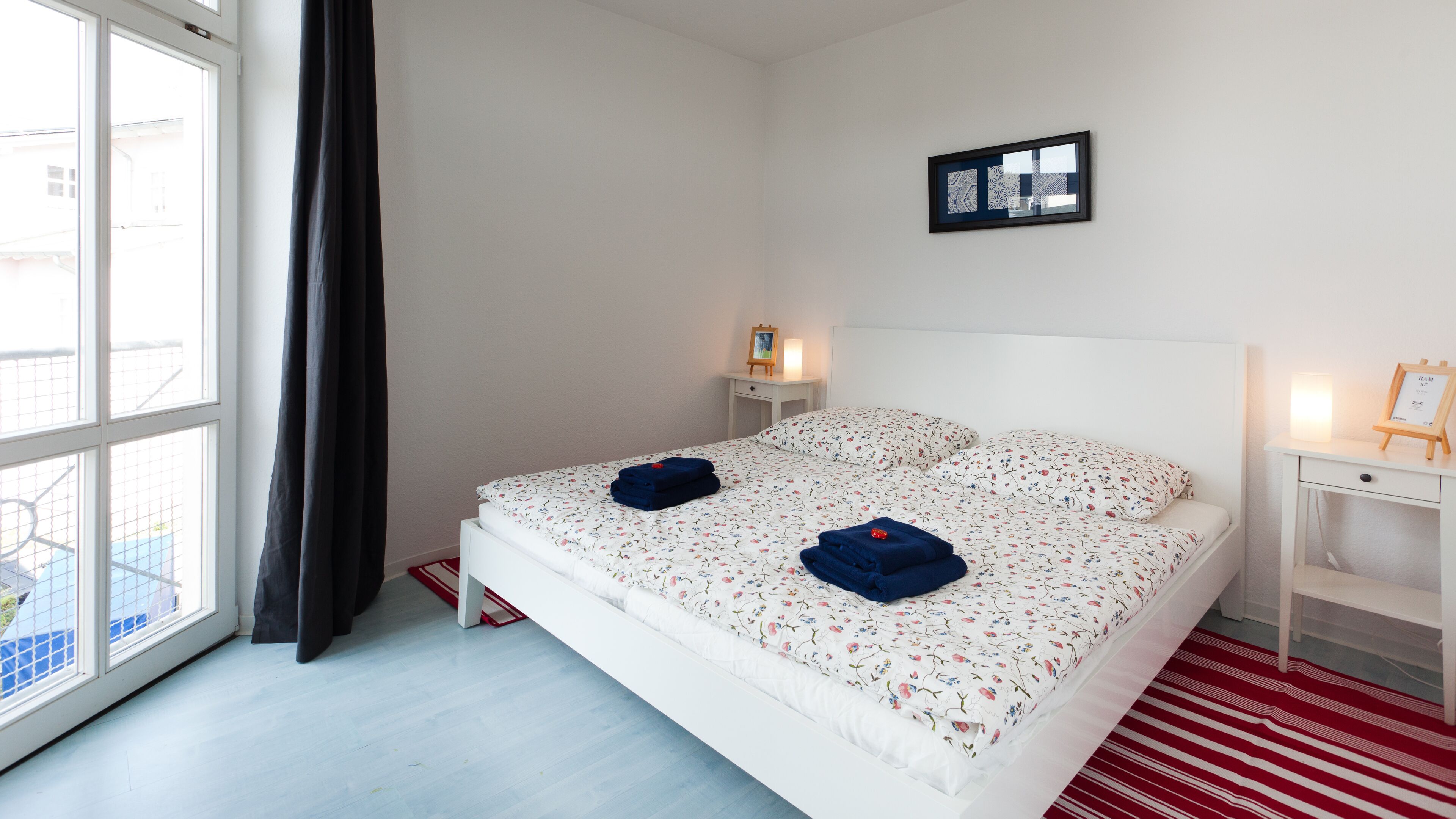 Ett hotellrum med en säng, ett sängbord, en liten avlastningsyta, ett fönster med gardiner och en tavla som hänger på väggen.