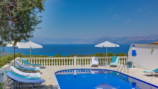 Zona junto a la piscina con sillones de descanso y sombrillas, con vistas panorámicas al mar y las montañas.