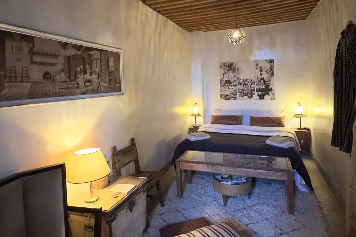 A cozy bedroom with a wooden ceiling, a bed with a headboard, a desk with a lamp, and a chair.