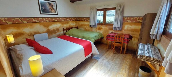 Un dormitorio con una cama, dos camas individuales, una mesa con una silla, una lámpara y una ventana con cortinas.
