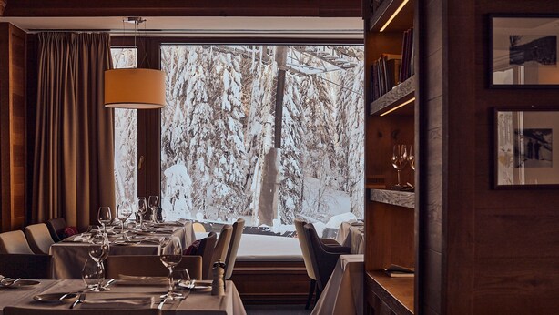 Un comedor con mesas dispuestas para una comida, un ventanal con vistas nevadas y una estantería con fotografías enmarcadas.