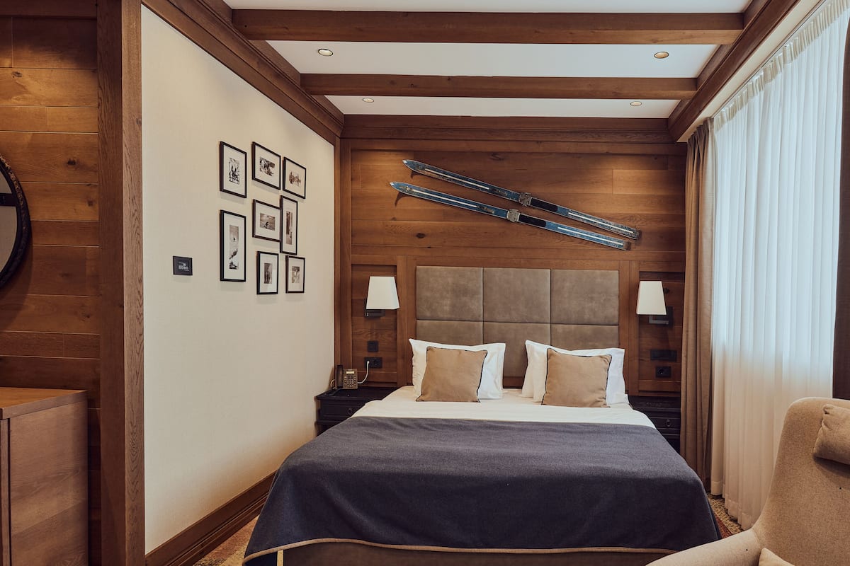 A hotel room with a wooden headboard, a bed with a dark blue bedspread, and a chair. There are framed pictures on the wall, a telephone on the nightstand, and a lamp.
