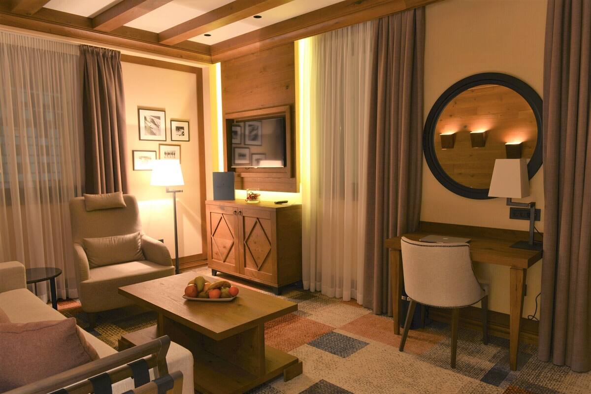 A hotel room with a sofa, armchair, wooden coffee table, desk, and a television.