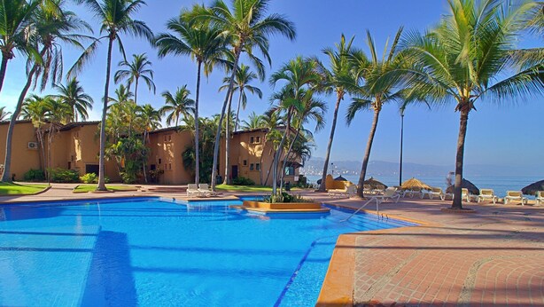 A swimming pool surrounded by palm trees and lounge chairs, with a view of the ocean and buildings in the background.
