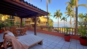 A balcony with a tiled floor, a covered seating area with chairs and a table, and potted plants.