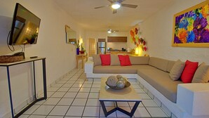 A modern living room with a sectional sofa, a coffee table, a flat-screen TV, and a kitchen in the background.