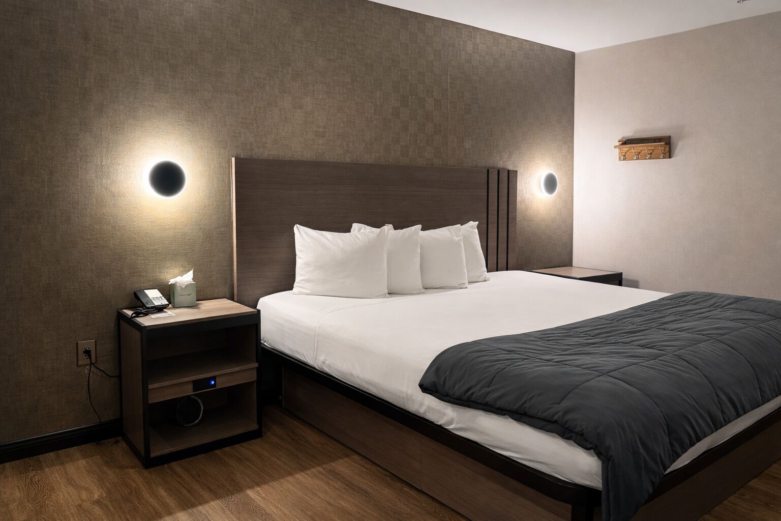 A modern hotel room with a large bed, a bedside table with a phone and tissues, and wall-mounted lighting.