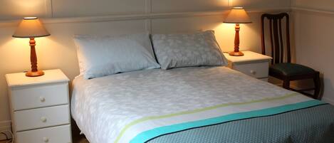 A neatly made bed with a patterned bedspread, two bedside tables with lamps, a chair, and a nightstand with drawers.