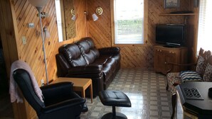 A cozy room with a brown leather sofa, a black office chair, a small wooden table, and a television on a wooden cabinet. - Schurmans Shore Cottage #4 (Summerside)
