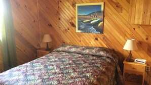 A wooden-paneled room with a bed, two bedside lamps, a nightstand with a lamp, and a framed picture on the wall. - Schurmans Shore Cottage #4 (Summerside)