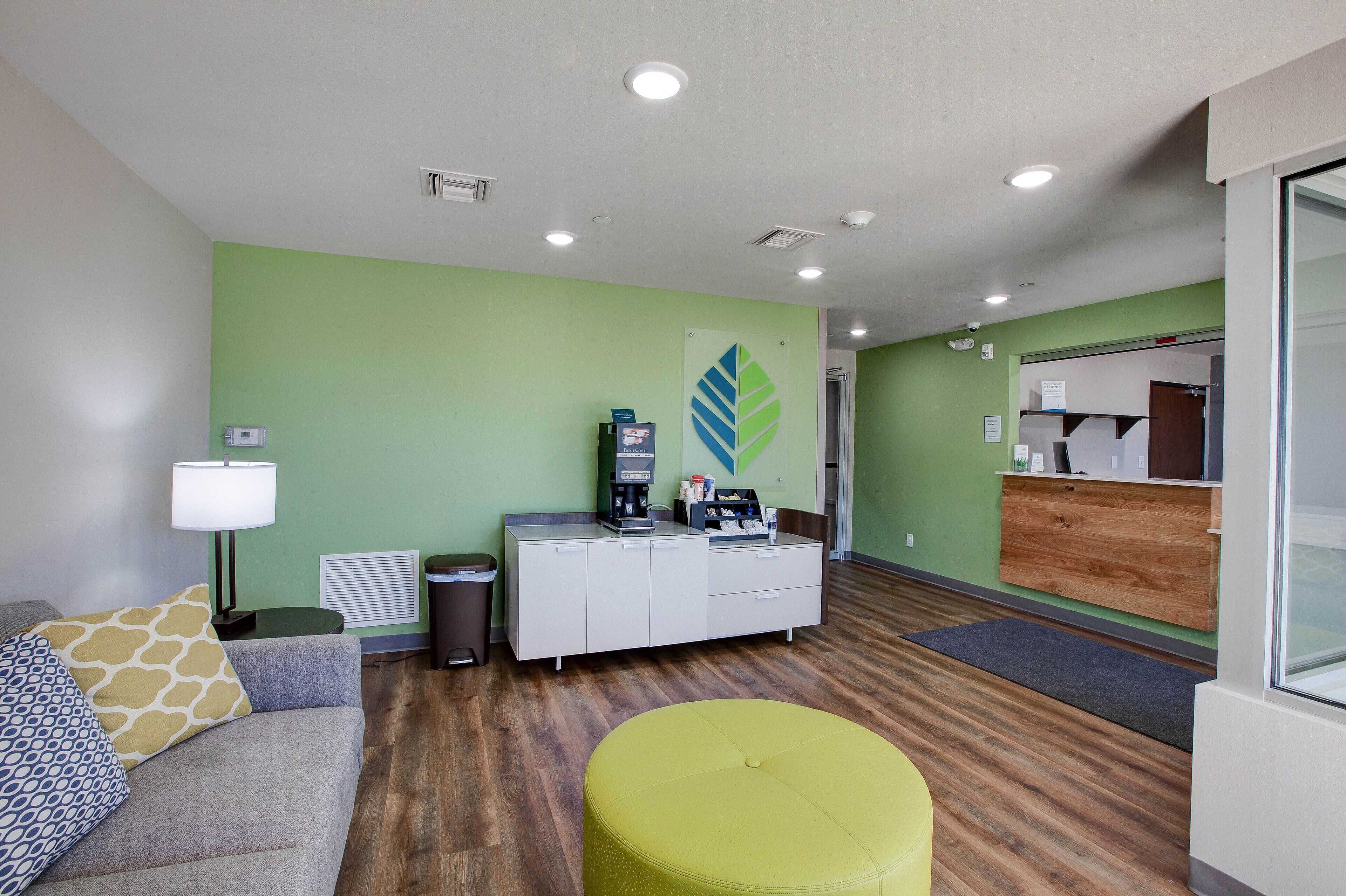A modern reception area with a green wall, a wooden reception desk, a coffee machine, and a seating area with a grey sofa and a yellow ottoman.