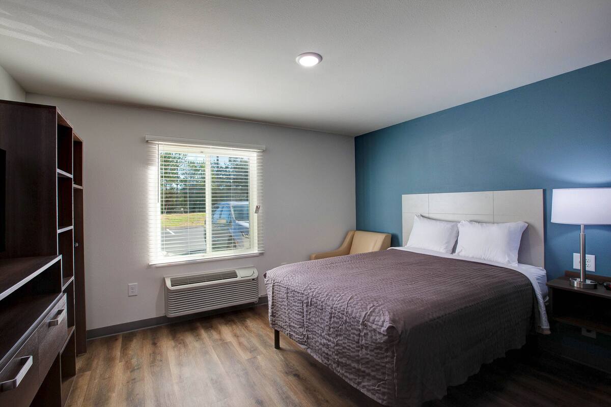 A bedroom with a bed, a chair, a desk, and a window with blinds.