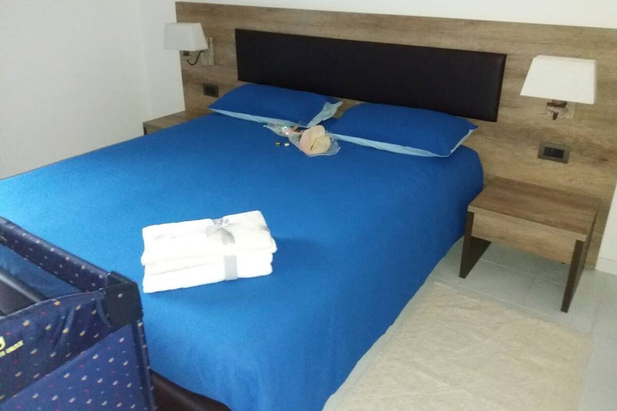 A neatly made bed with a blue bedspread and pillows, a wooden headboard, bedside lamps, and a small wooden nightstand.