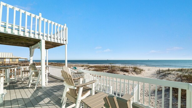 Une terrasse en bois, dotée de rampes blanches et de chaises de plage, offre une vue sur une plage de sable et l’océan.
