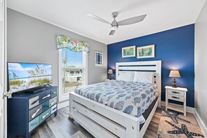 A bedroom with a bed, a TV, a ceiling fan, a window with blinds, and artwork on the walls. - Recently updated, beautifully furnished gulf front home with decks galore! (Gulf Shores)