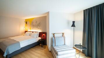 Habitación de hotel con cama, mesita de noche, lámpara, un taburete pequeño y una obra de arte en la pared.