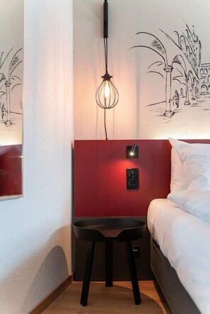 A hotel room with a bed, a nightstand, a desk with a chair, a wall-mounted light fixture, and a wall art depicting a building and palm trees. - Hotel Stockalperhof (Brig)