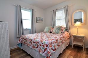 A bedroom with a bed, a bedside lamp, a wicker chair, and a wall-mounted mirror.
