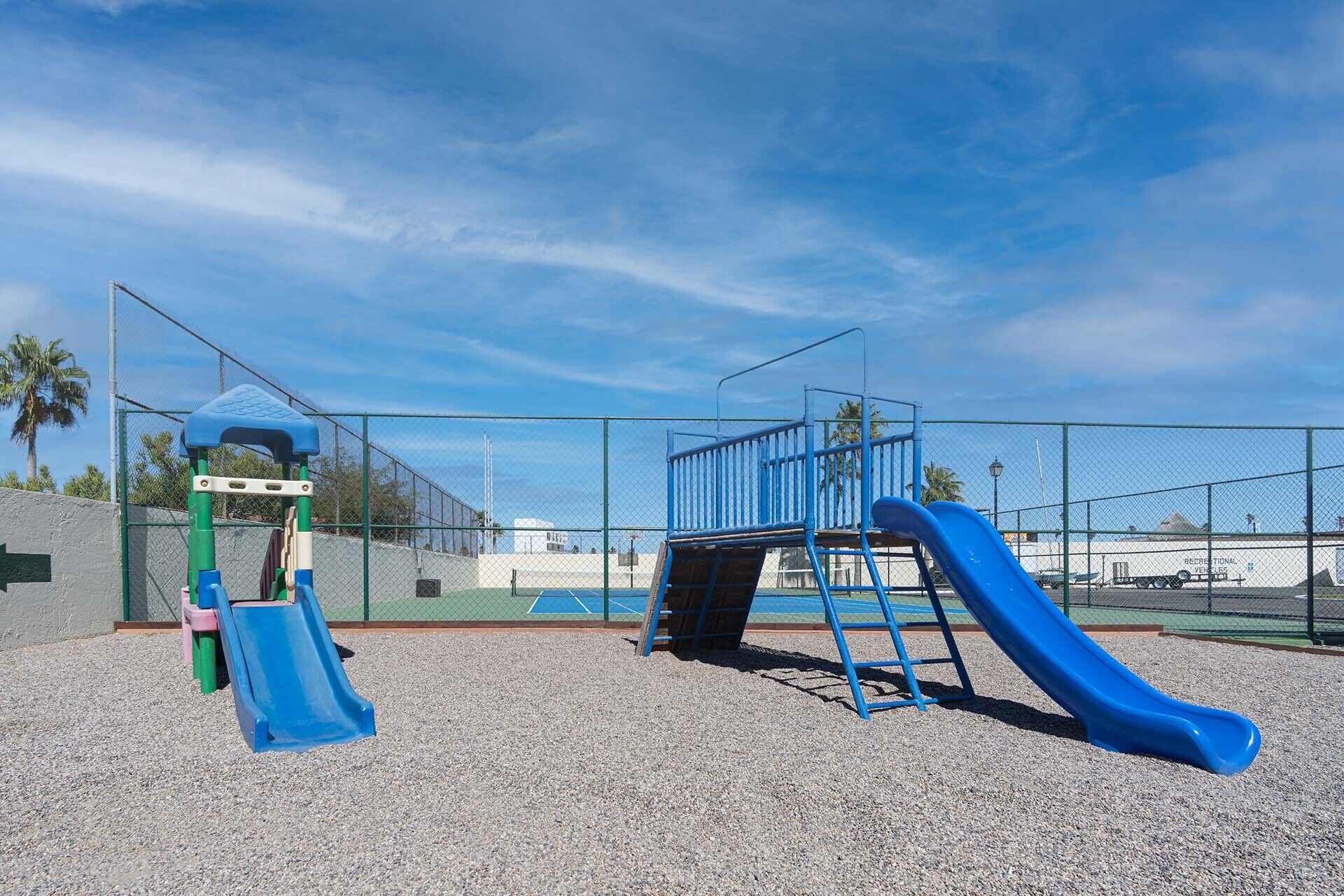 A playground with a blue slide and climbing structures, surrounded by a fence and palm trees.