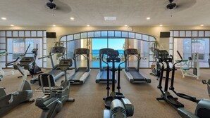A gym with treadmills, elliptical machines, and stationary bikes. It has large windows with a view of the sea.