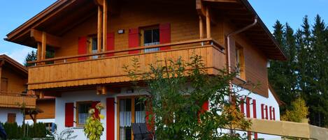 A two-story wooden chalet with a balcony, red shutters, and a white facade.
