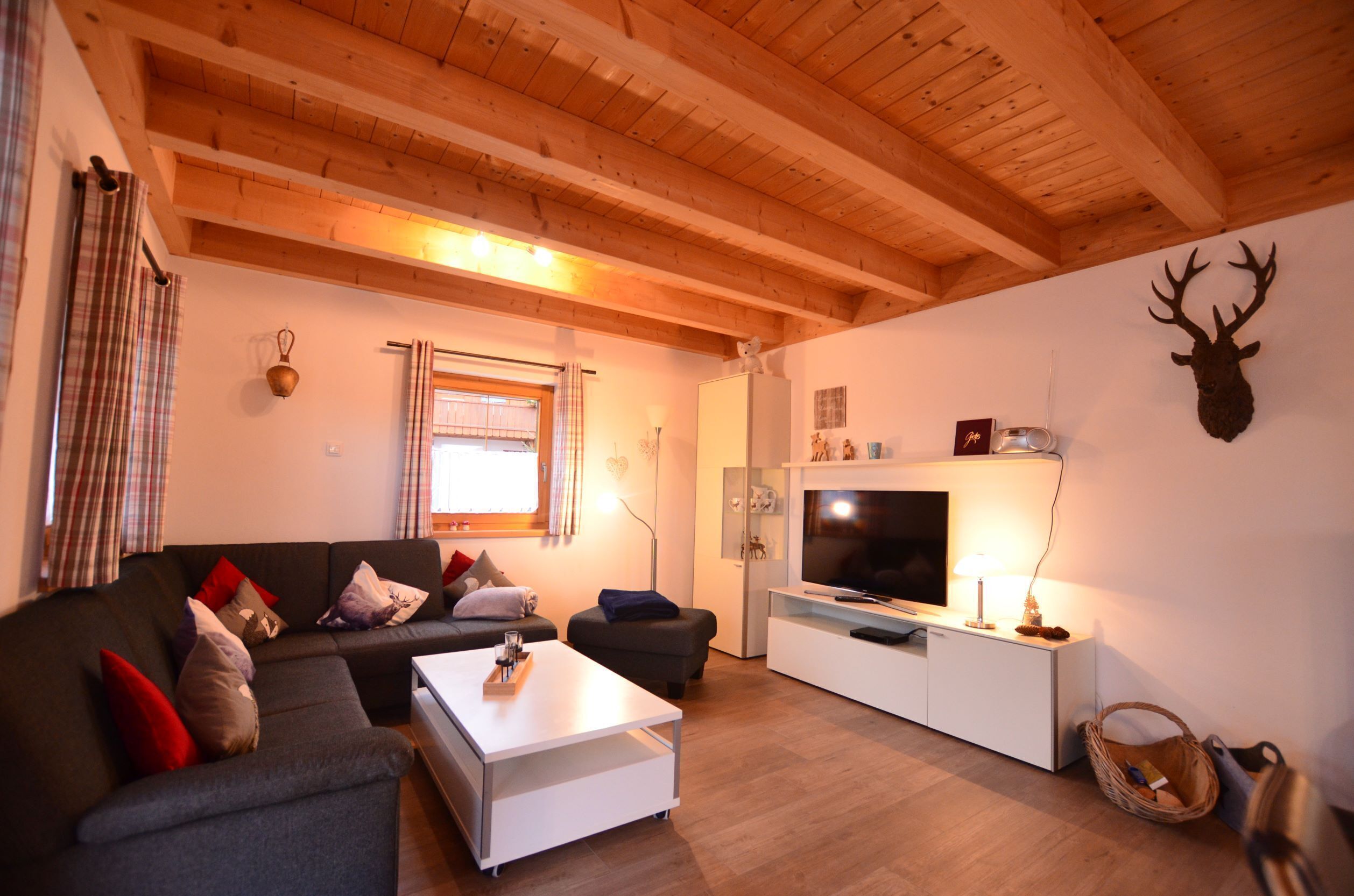 A modern living room with a wooden ceiling, a grey sofa, a white coffee table, a flat-screen TV, and a mounted deer head on the wall.
