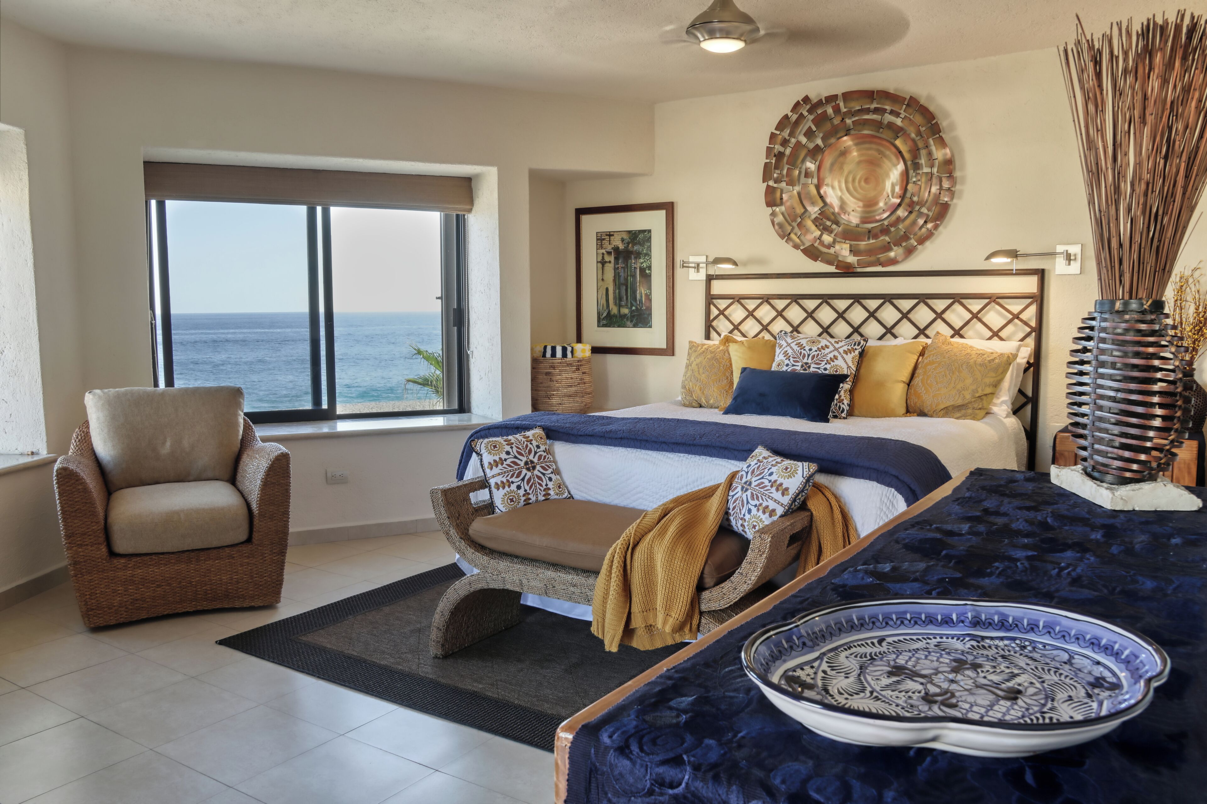 A bedroom with a large bed, a chair, a view of the sea, and a decorative wall piece.