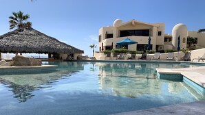 A swimming pool with lounge chairs and umbrellas, a thatched roof structure, and a building with a unique architectural design. - Beach front 2 Bedroom 2 Bathroom luxury condo with amazing views.  (Cabo San Lucas)