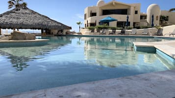 A swimming pool with lounge chairs and umbrellas, a thatched roof structure, and a building with a unique architectural design.