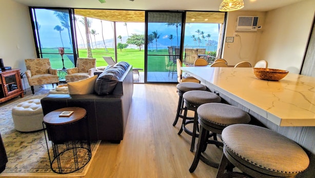 Un salon moderne doté d’un grand téléviseur, de sièges confortables, d’un coin cuisine avec des tabourets de bar et d’une vue sur un terrain de golf grâce à de grandes fenêtres.