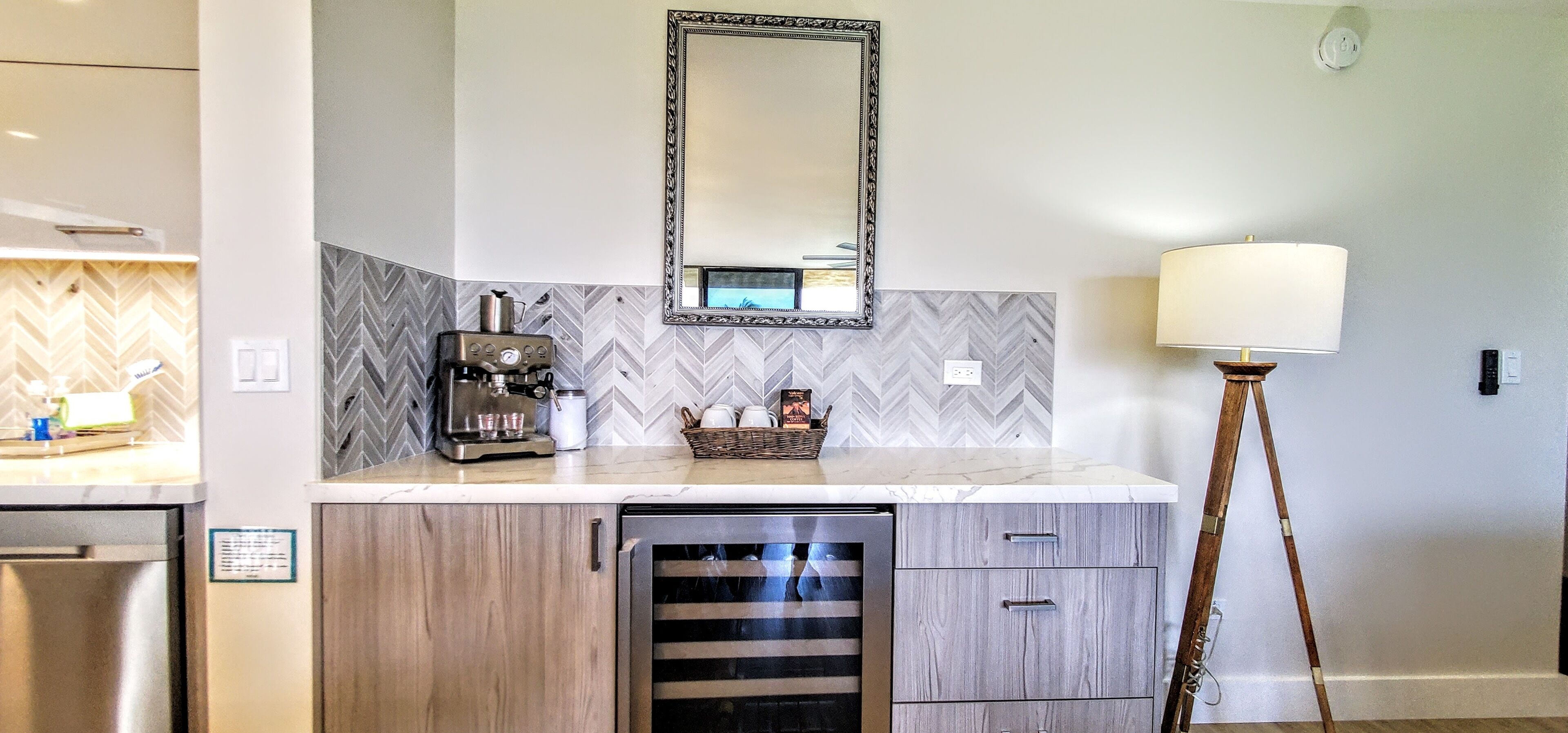 A modern kitchen with a coffee machine, a microwave, and a dishwasher. There is a mirror above the counter and a floor lamp to the right.