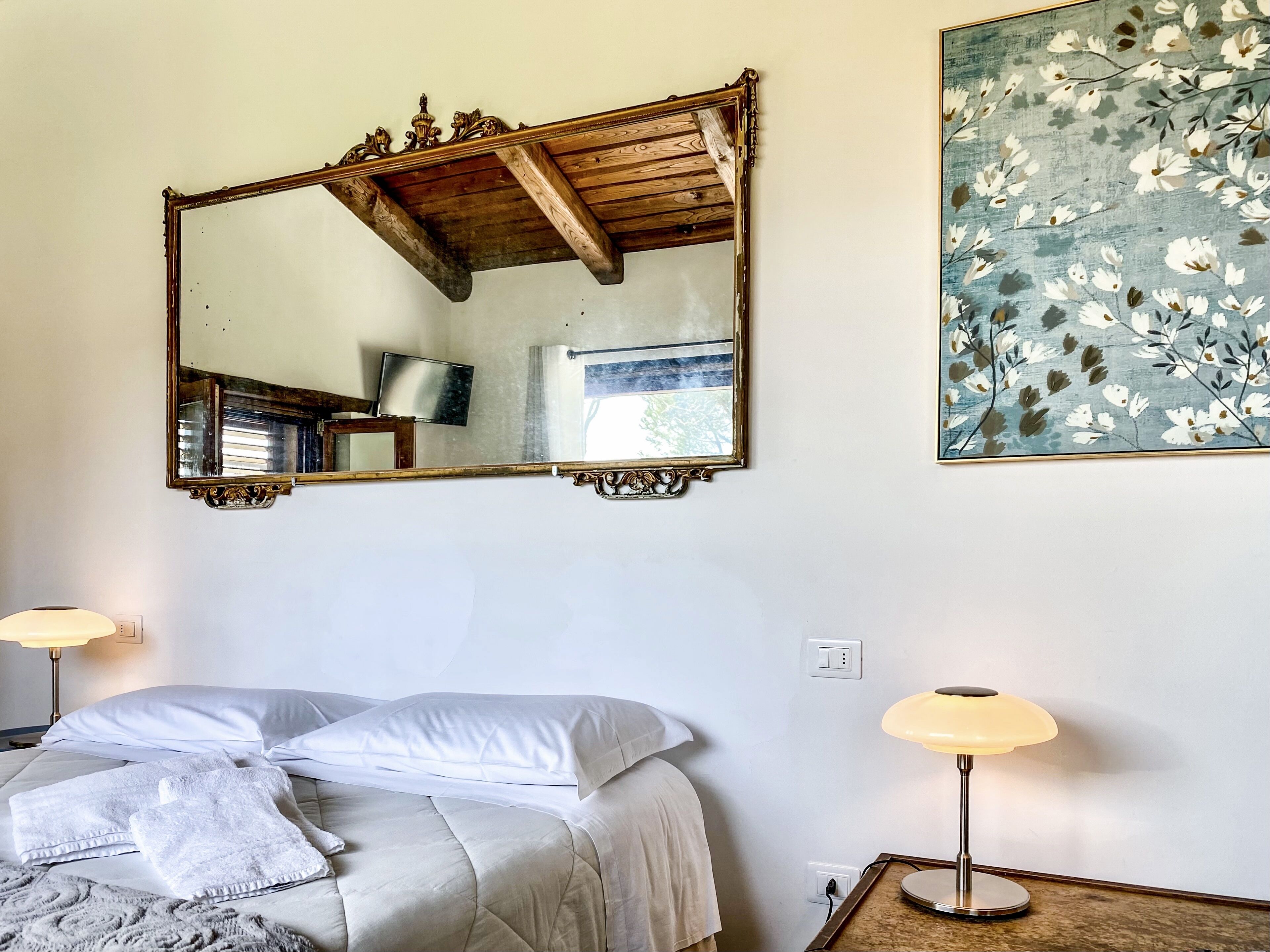 A bedroom with a bed, two bedside lamps, a large mirror, and a floral painting on the wall.