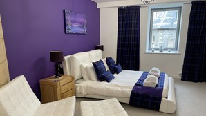 Un dormitorio con una cama, dos sillas blancas, una mesita de noche, una lámpara y una ventana con cortinas.