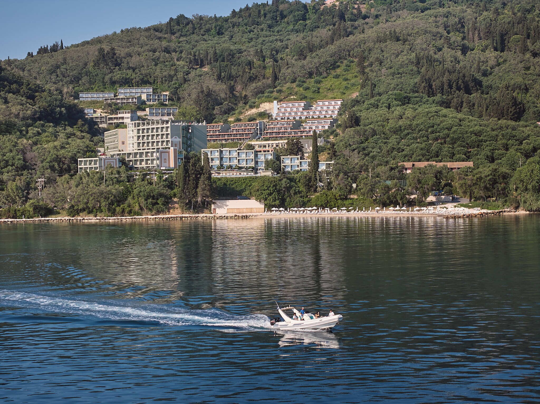 Um hotel na orla marítima com vários edifícios, um barco na água e um cenário de colina ao fundo.