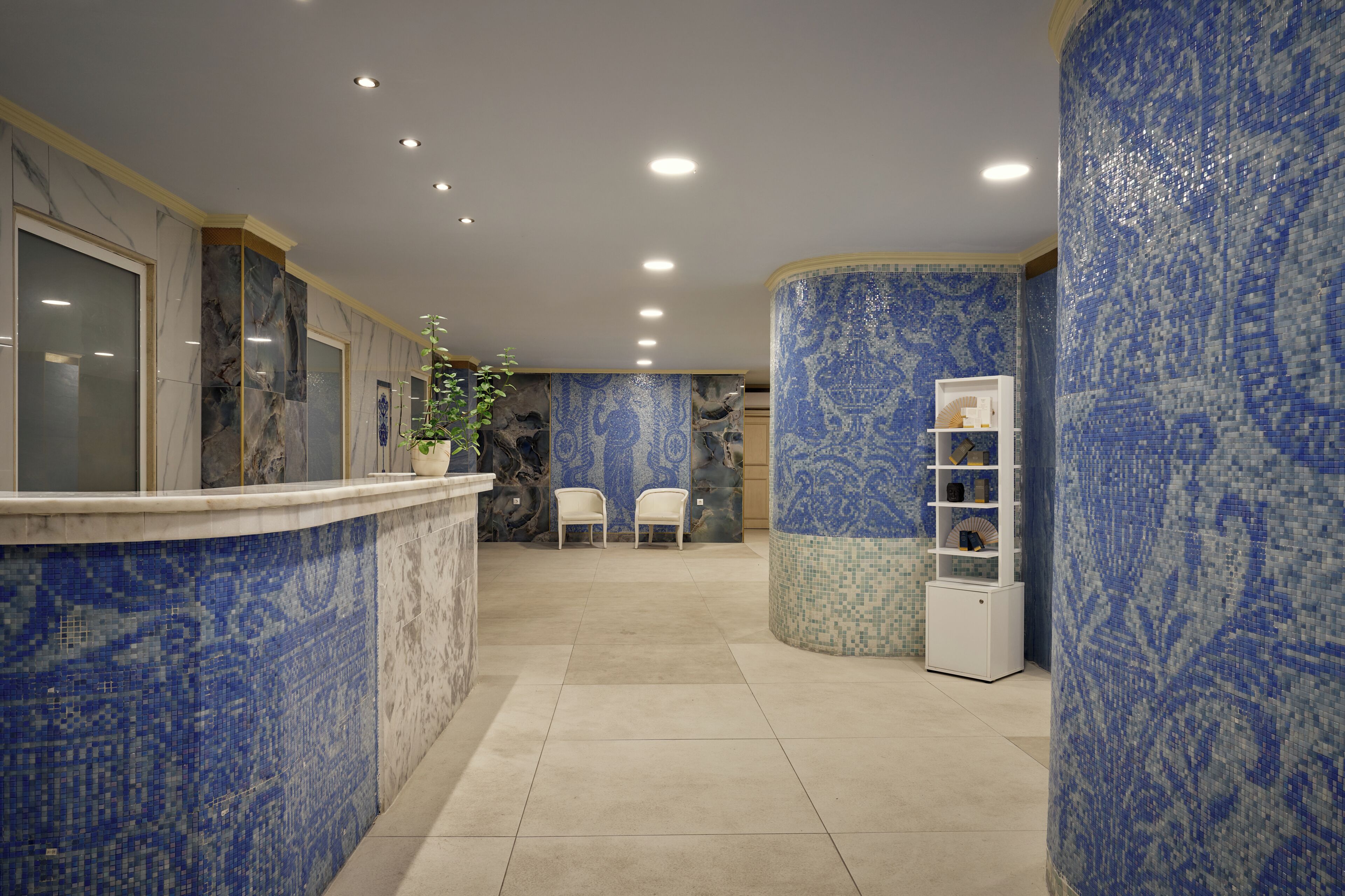 A reception area with a marble counter, two white chairs, a potted plant, and a decorative wall with blue mosaic tiles.