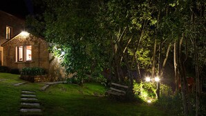 Een stenen huis met een goed verlichte tuin 's nachts, met een houten bankje en een stenen pad dat naar het huis leidt.