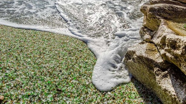 Een strand met helder water, kiezelstenen en een rotsachtige richel.