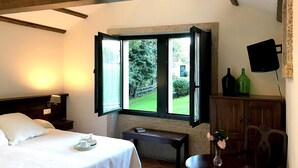 Een slaapkamer met een bed, een nachtkastje, een tafel met een vaas en een raam met een open luik.