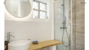 Une salle de bain avec un lavabo blanc, un miroir rond, une fenêtre ornée d’un motif décoratif et un espace douche doté d’une porte en verre.