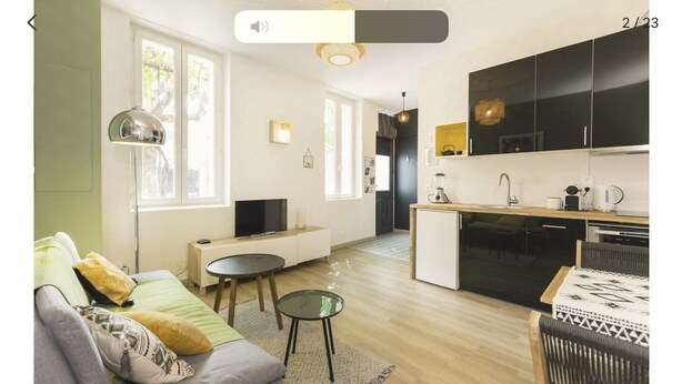 Un salon moderne avec un canapé, une table basse et un téléviseur. On peut apercevoir une cuisine avec des meubles noirs et un coin repas comprenant une table et des chaises.