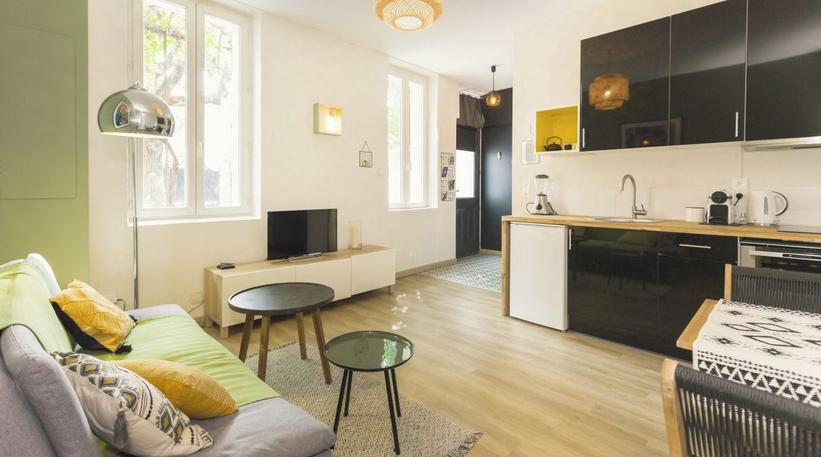 A modern living room with a sofa, coffee table, and TV. A kitchen with black cabinets and a dining area with a table and chairs is visible.