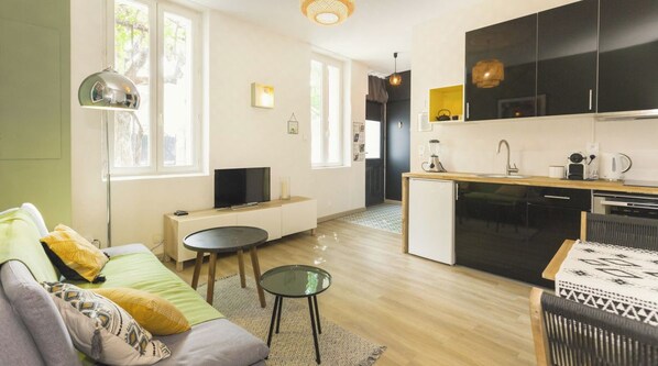 Un salon moderne avec un canapé, une table basse et un téléviseur. On peut apercevoir une cuisine avec des meubles noirs et un coin repas comprenant une table et des chaises.