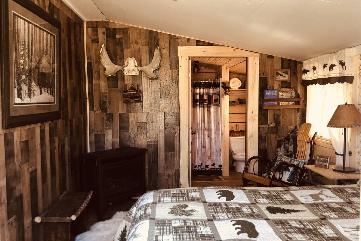 Una habitación rústica con pared de madera, una cama con colcha de estampados animales, chimenea y una puerta que da al baño.