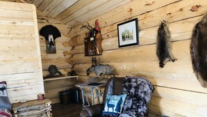 A rustic cabin interior with a leather armchair, wooden shelves, and various decorative items including animal pelts and a framed picture. - Wrangell Mountain Lodge (Chitina)