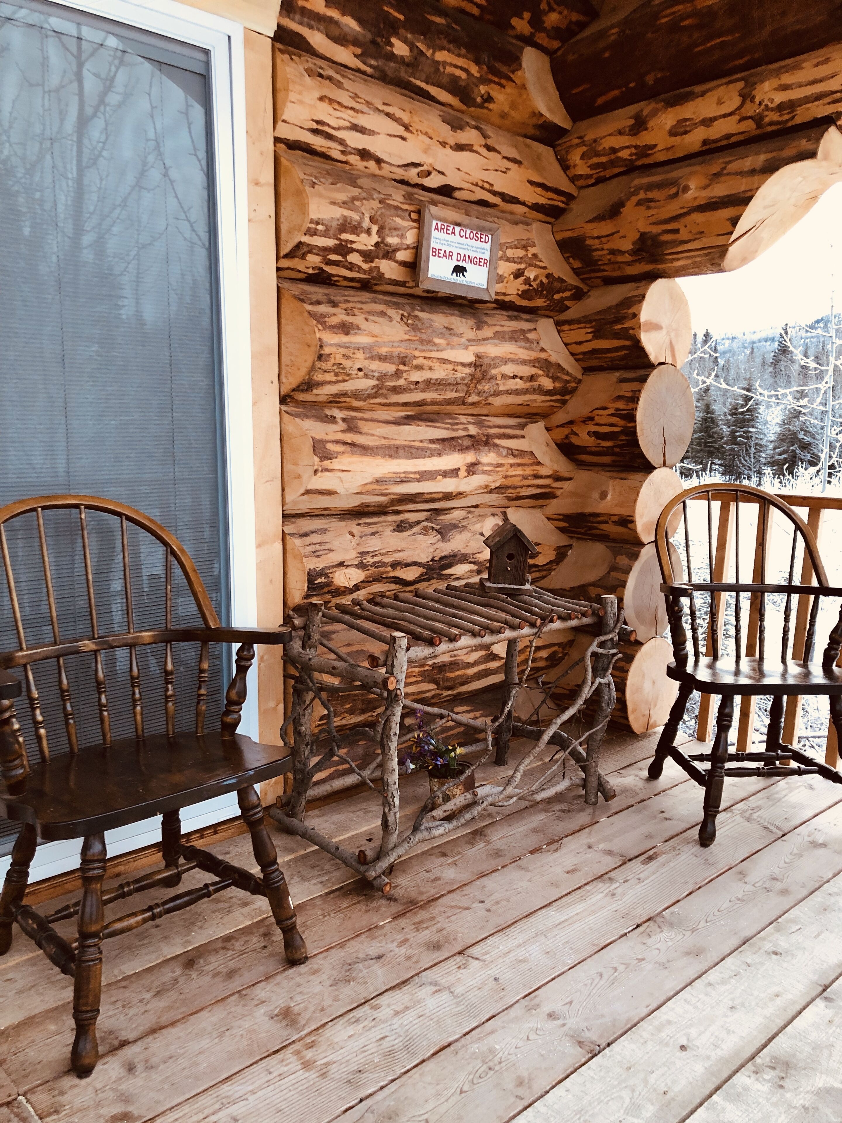 A rustic wooden cabin with a bear danger sign, two wooden chairs, and a birdhouse on the porch.